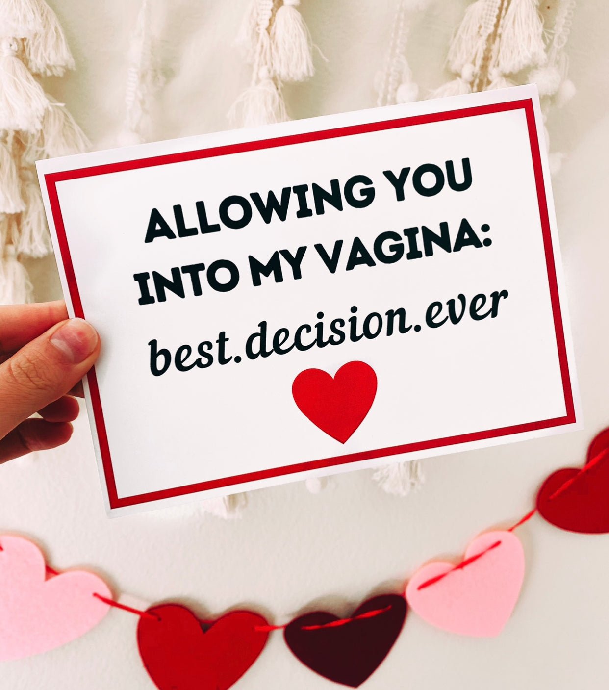 A hand holding a white greeting card with a red border and black text that reads “Allowing you into my vagina: best decision ever,” with a red heart printed below the text. The background includes hanging tassels and a garland of red and pink heart shapes.