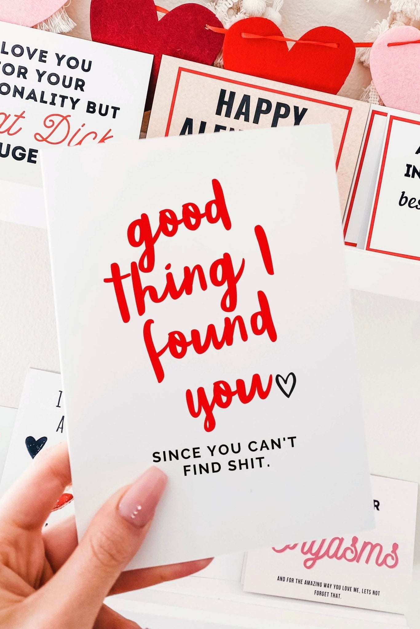 Greeting card with red and black text on a white background, photographed being held by a hand with other Valentine themed cards visible in the background
