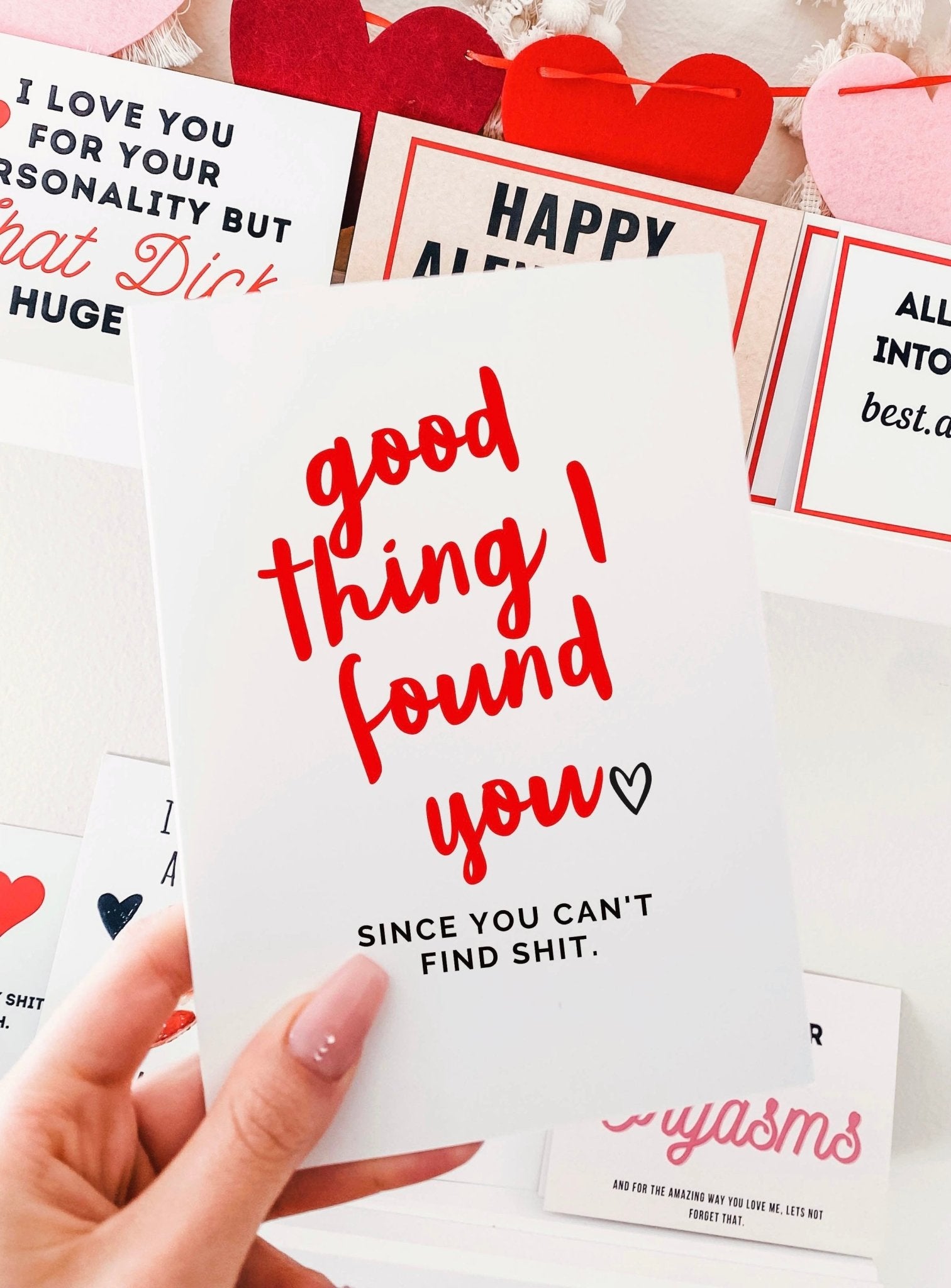 Greeting card with red and black text on a white background, photographed being held by a hand with other Valentine themed cards visible in the background