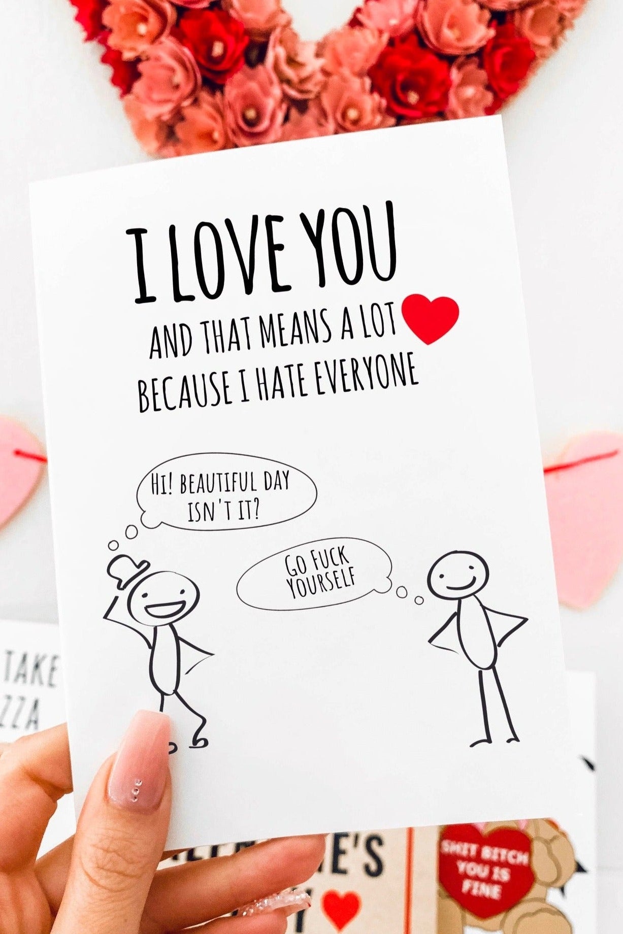 Greeting card with black text and a small red heart above simple cartoon figures, photographed being held by a hand with Valentine themed decorations in the background