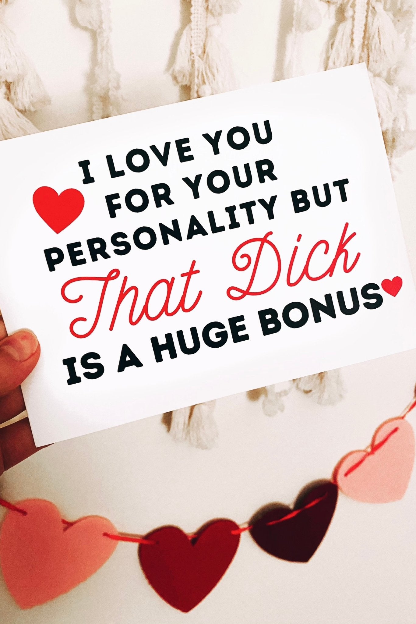 Greeting card displaying a humorous adult phrase in black and red text with heart graphics, photographed being held by a hand against a white background with white fring and hearts