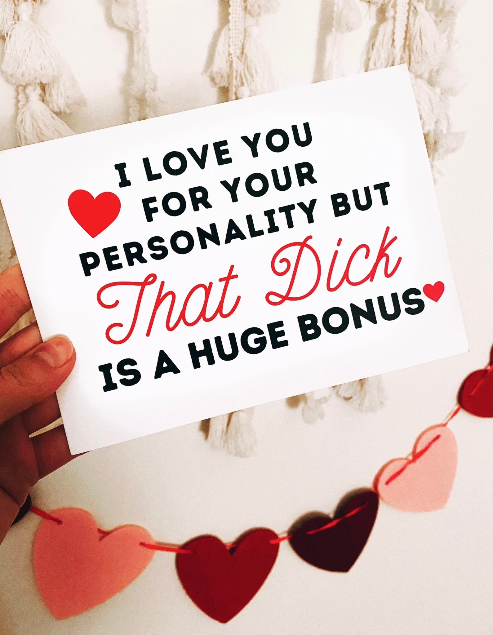 Greeting card displaying a humorous adult phrase in black and red text with heart graphics, photographed being held by a hand against a white background with white fring and hearts