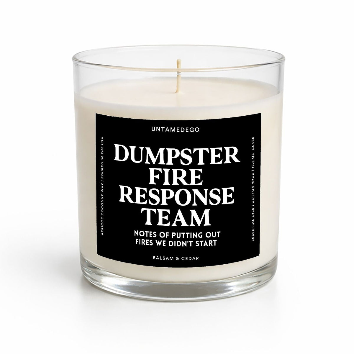 A clear glass jar candle with cream-colored wax and a black label that reads 'DUMPSTER FIRE RESPONSE TEAM' in white text, photographed on a clean white background.