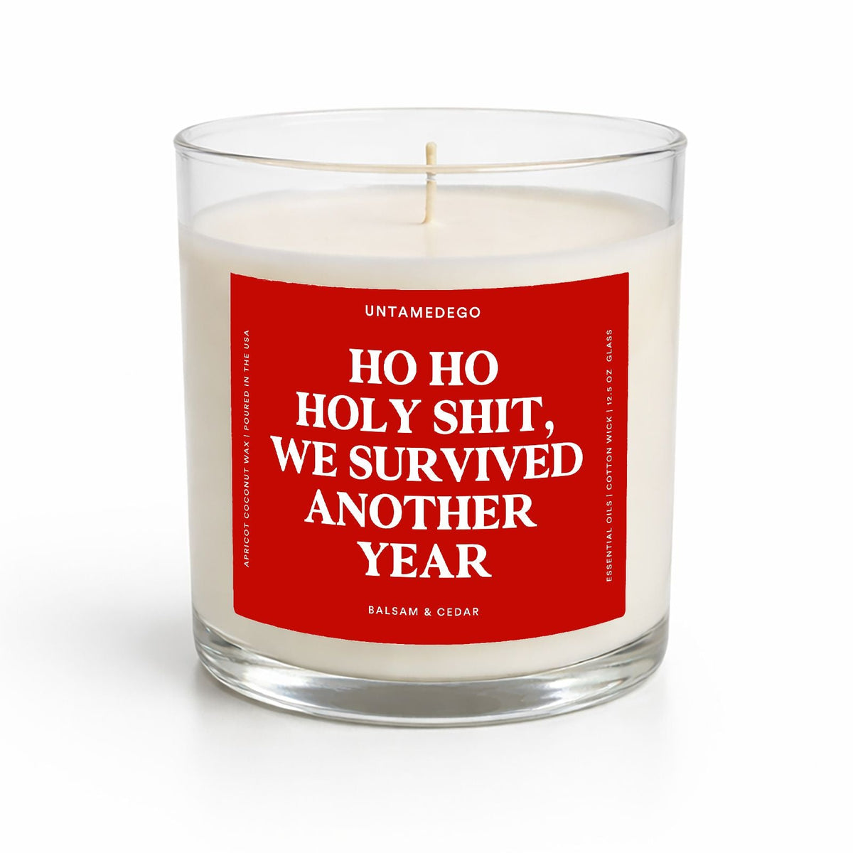 A clear glass jar candle with white wax and a bold red front label reading 'HO HO HOLY SHIT, WE SURVIVED ANOTHER YEAR' in white text. The brand name UNTAMEDEO is visible at the top of the label, with 'BALSAM & CEDAR' at the bottom.
