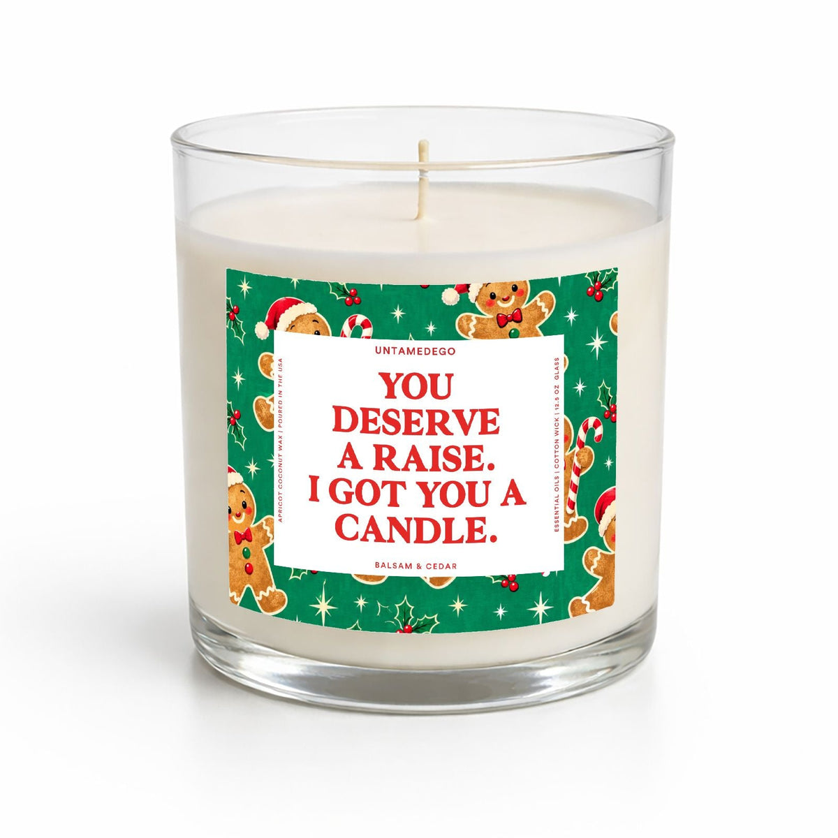 A clear glass jar candle with white wax and a single wick, featuring a festive green holiday label with gingerbread illustrations and the text 'YOU DESERVE A RAISE, I GOT YOU A CANDLE.' on a white background.