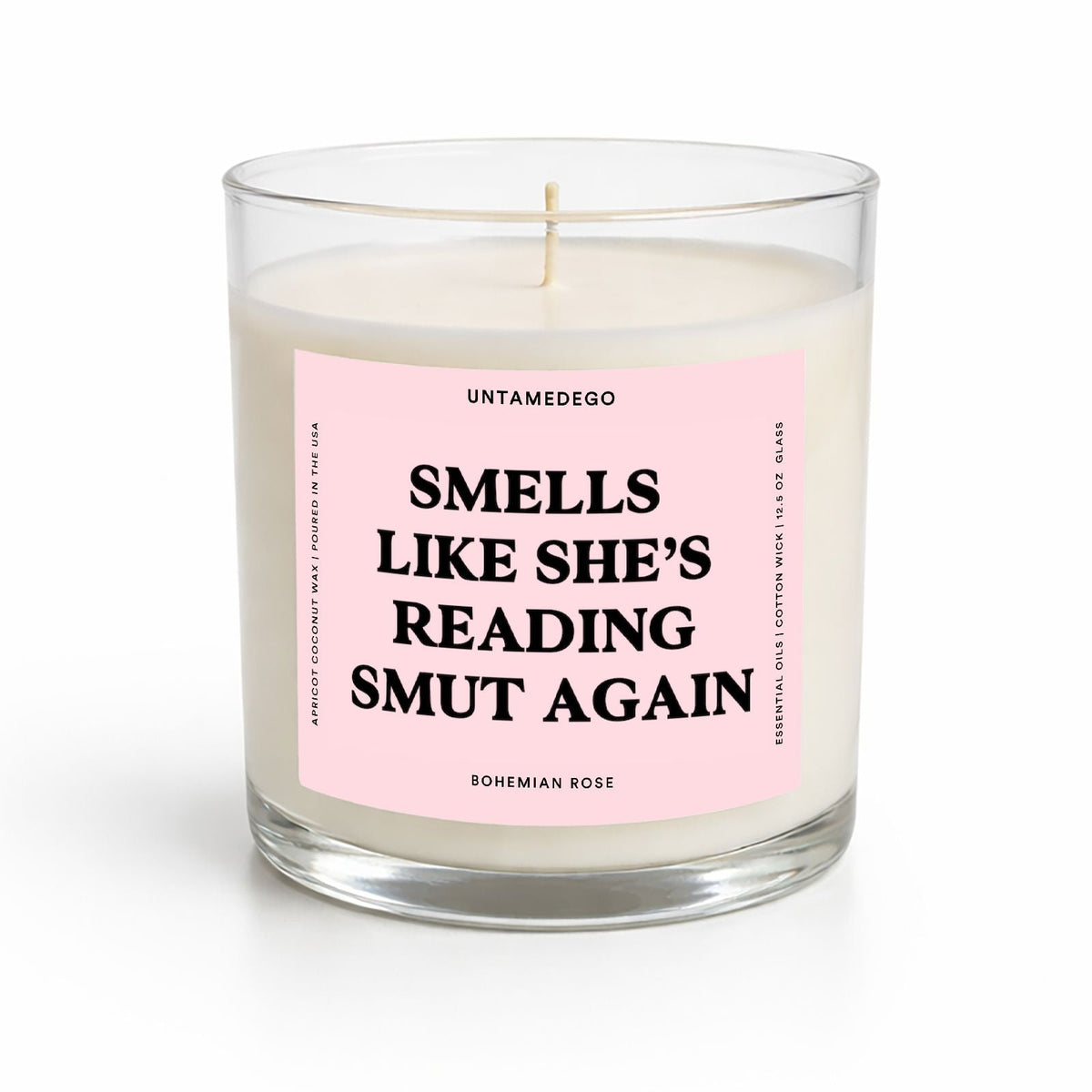 A glass jar candle with white wax and a pink label that reads 'SMELLS LIKE SHE'S READING SMUT AGAIN' by Untamedgo, on a clean white background.