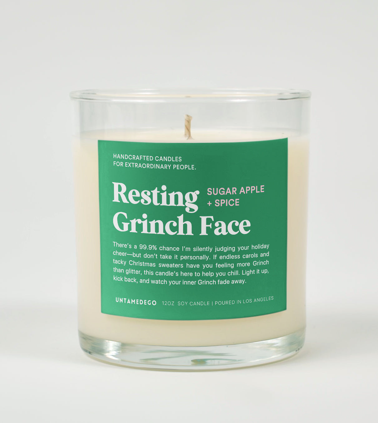 A clear glass candle with a label reading 'Resting Grinch Face' with additional text mentioning 'Sugar Apple Spice'. The candle is placed in a clear glass with a lid.