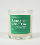 A clear glass candle with a label reading 'Resting Grinch Face' with additional text mentioning 'Sugar Apple Spice'. The candle is placed in a clear glass with a lid.