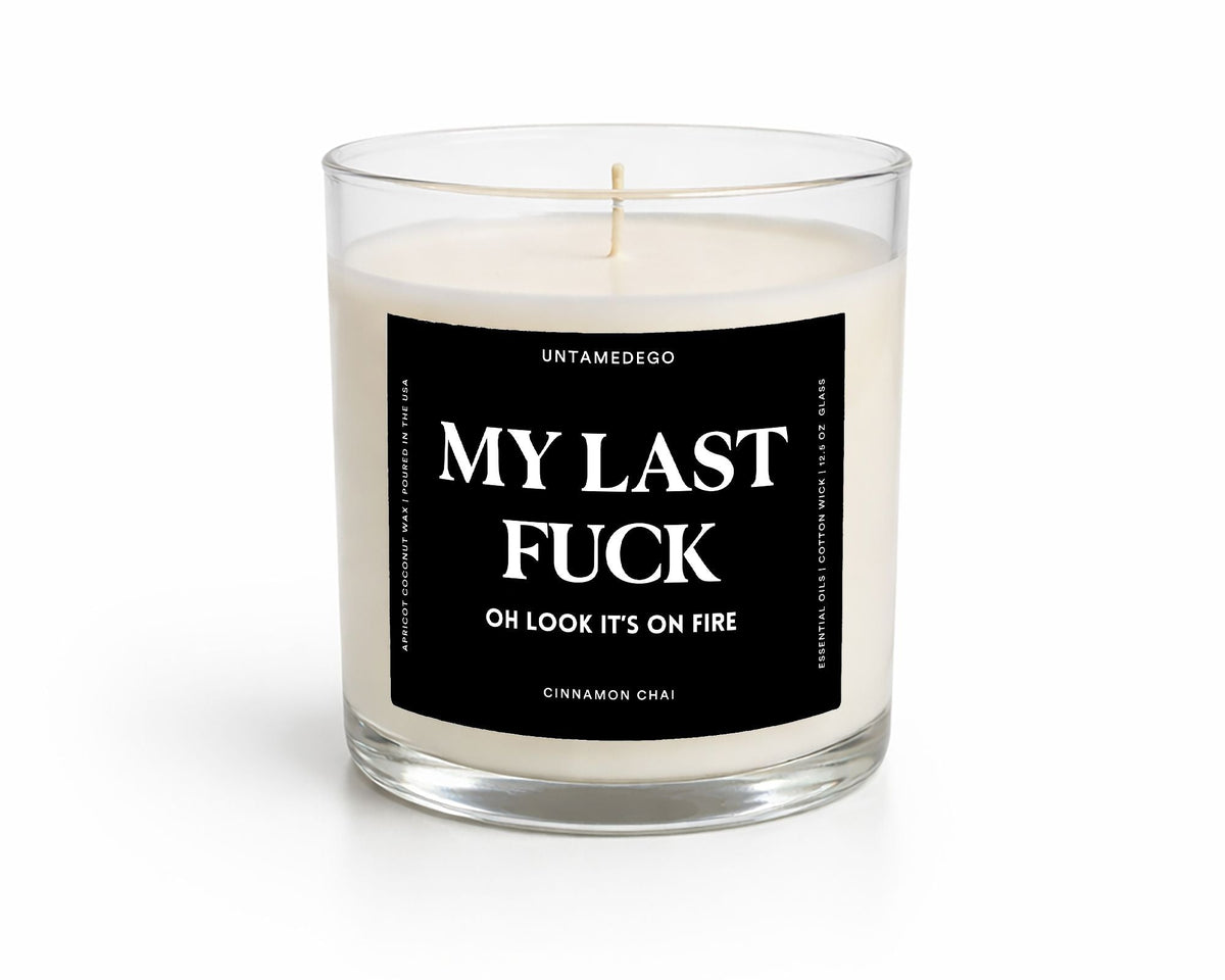 A white soy wax candle in a clear glass jar with a black square label that reads 'MY LAST FUCK OH LOOK IT'S ON FIRE' and smaller text 'UNTAMEDEGO' and 'COASTAL AMBER | CEDAR'.