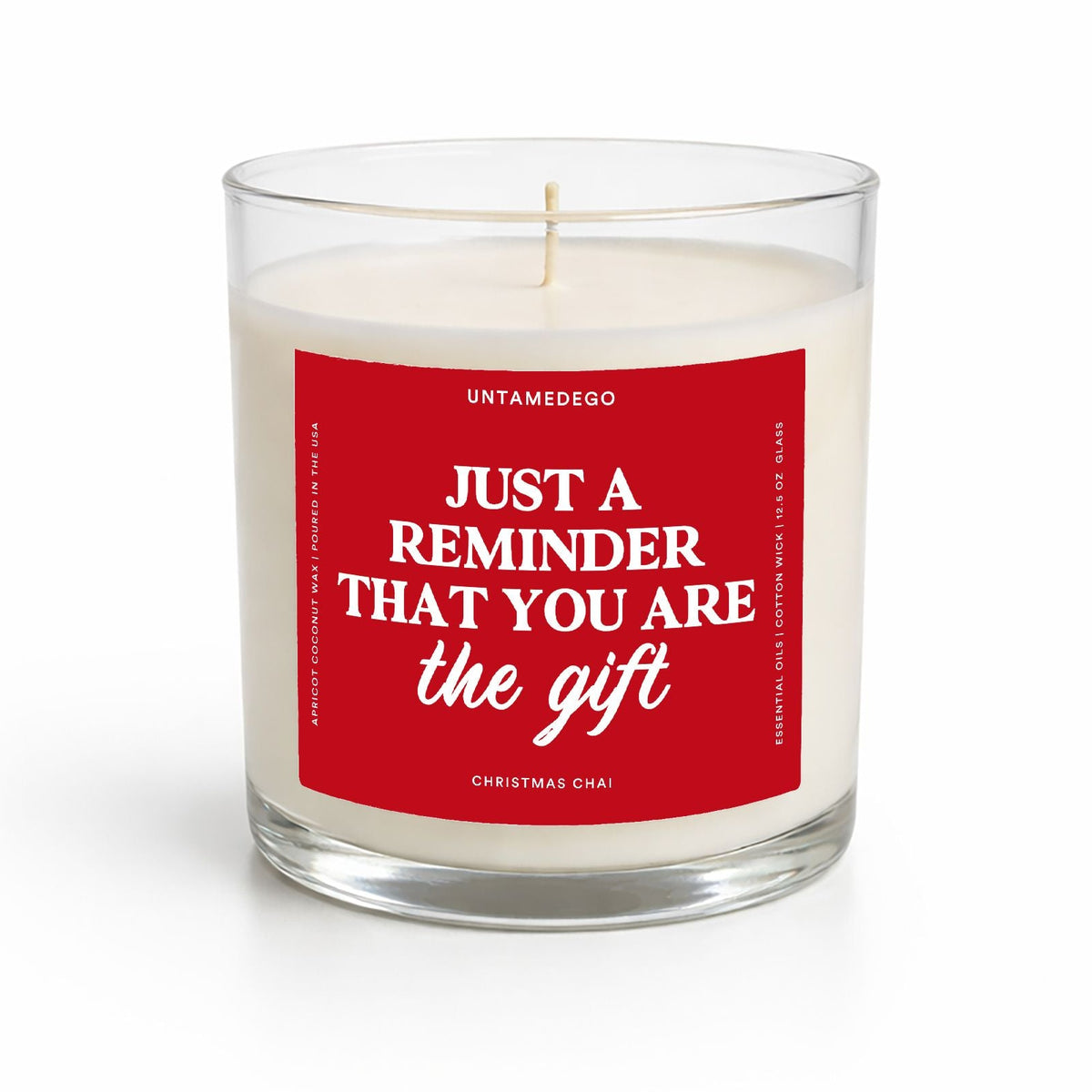 A white soy candle in a clear glass jar with a red square label that reads, 'JUST A REMINDER THAT YOU ARE the gift' in white text. The brand name 'UNTAMEDEGO' is visible at the top of the label.