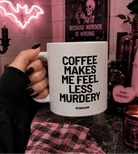 A hand with black nail polish holds a white ceramic mug printed with the text 'COFFEE MAKES ME FEEL LESS MURDERY' and 'UNETHICALGAY'. The background is a dimly lit, gothic-themed room with pink ambient lighting, a pink Smeg kitchen appliance, a pink heart-shaped candle, and a pink plaid cloth.