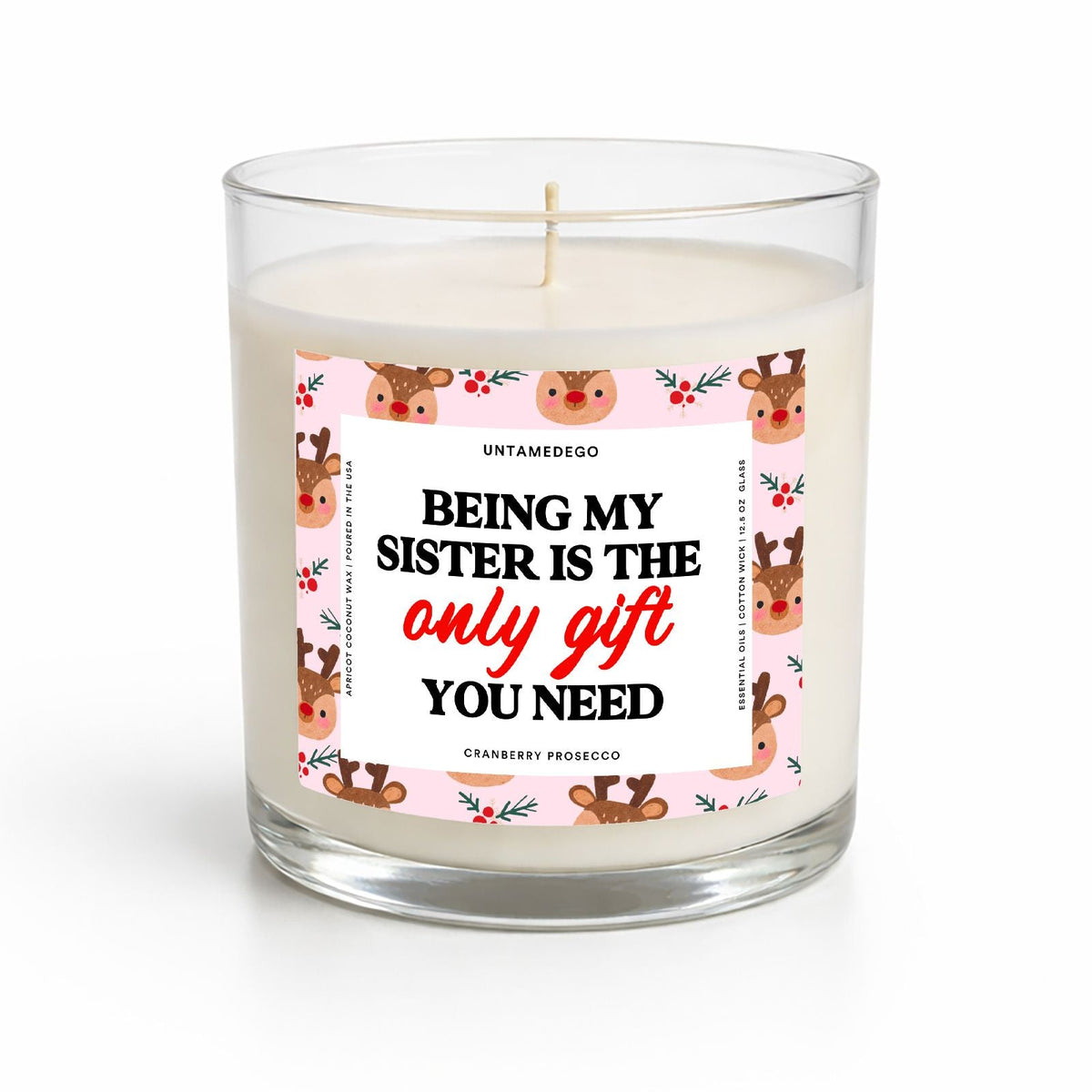 A 16 oz scented candle in a clear glass jar on a white background. The candle has cream-colored wax and a white paper label featuring a pink border with cartoon reindeer faces and the text 'BEING MY SISTER IS THE only gift YOU NEED' in black and red lettering.