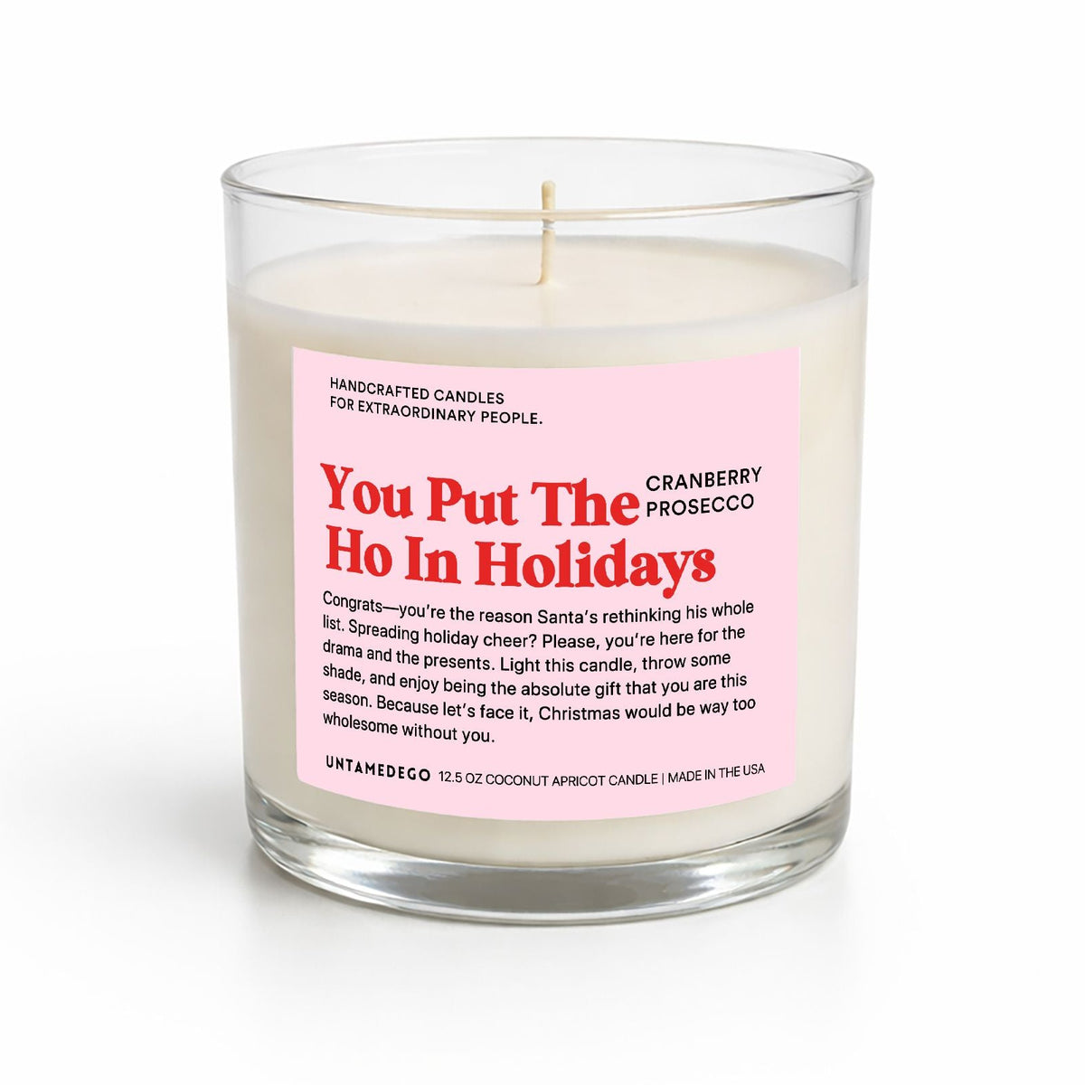A handcrafted coconut soy candle in a clear glass jar with a pink and red label that reads 'You Put The Ho In Holidays' and 'Cranberry Prosecco'.