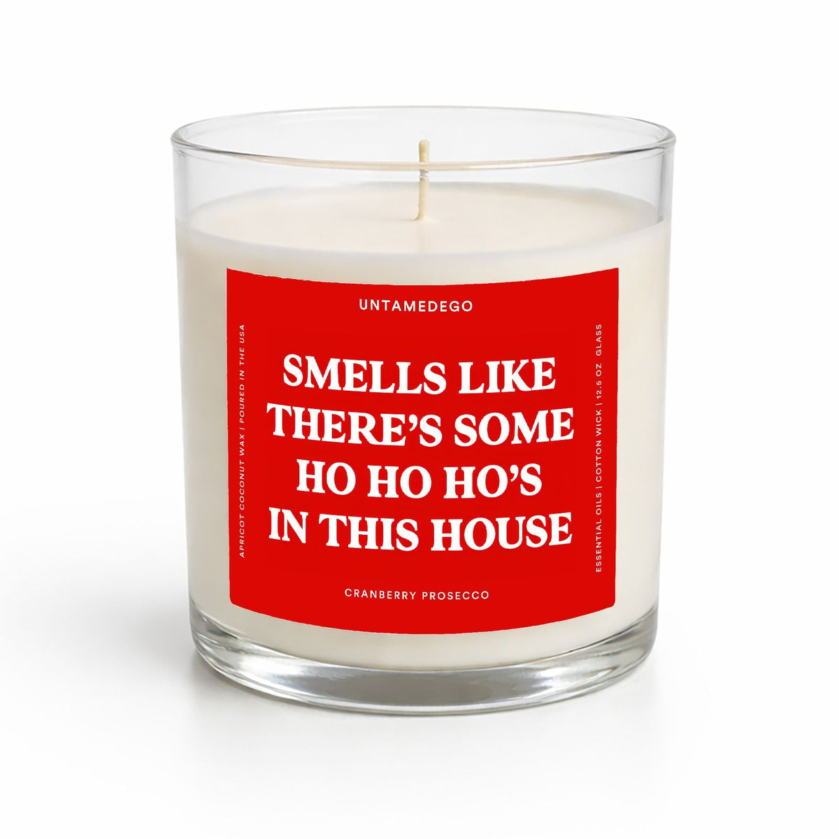 A white wax candle in a clear glass jar with a red front label that reads 'SMELLS LIKE THERE'S SOME HO HO HO'S IN THIS HOUSE' and smaller text 'UNTAMEDEGO' and 'CRANBERRY PROSECCO'.