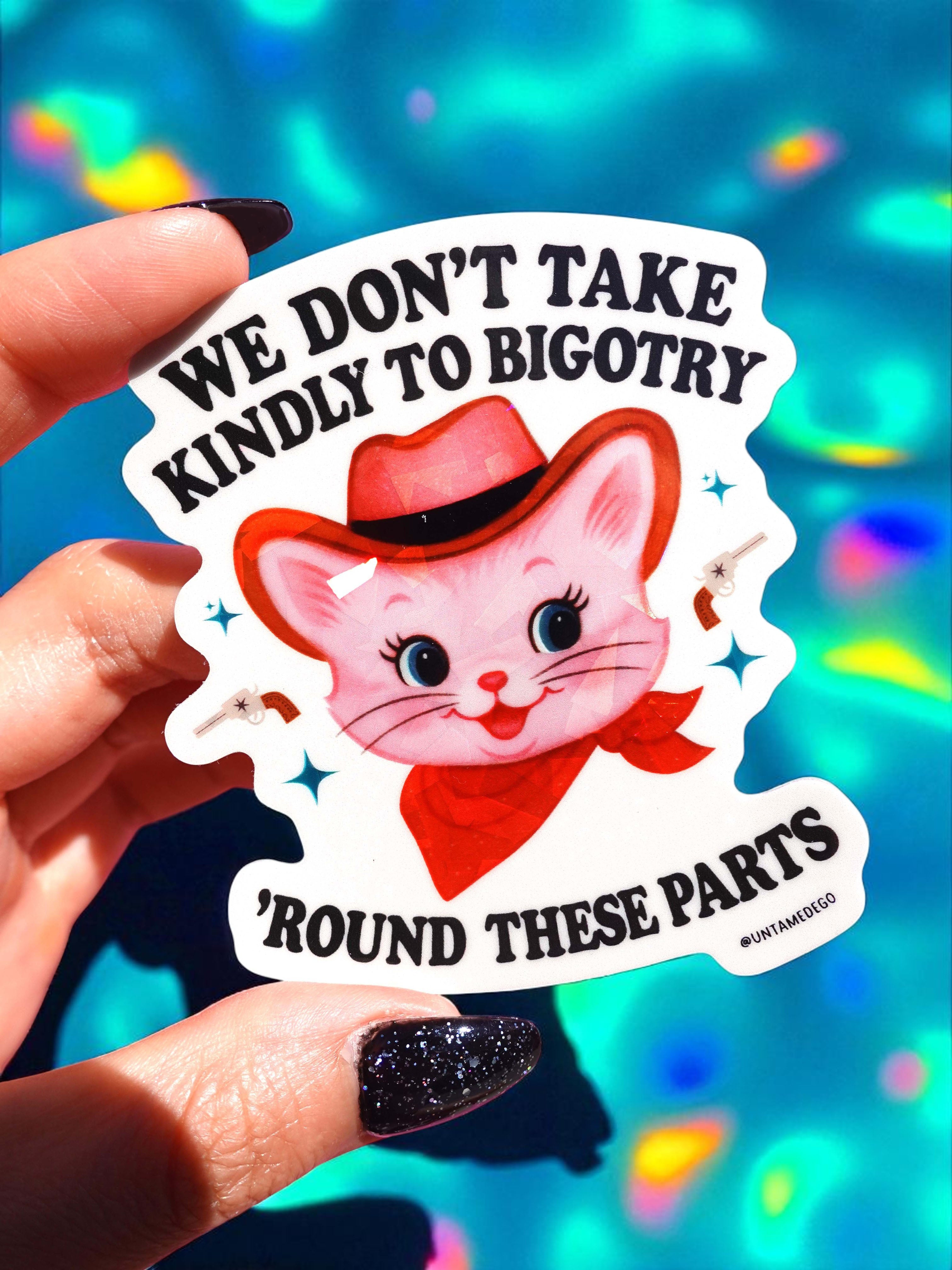Sticker with a pink cat wearing a cowboy hat and text about not taking kindly to bigotry, held against a colorful background.