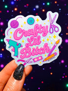 Sticker with 'Crafty Lil Bitch' text held by a hand against a colorful, starry background.