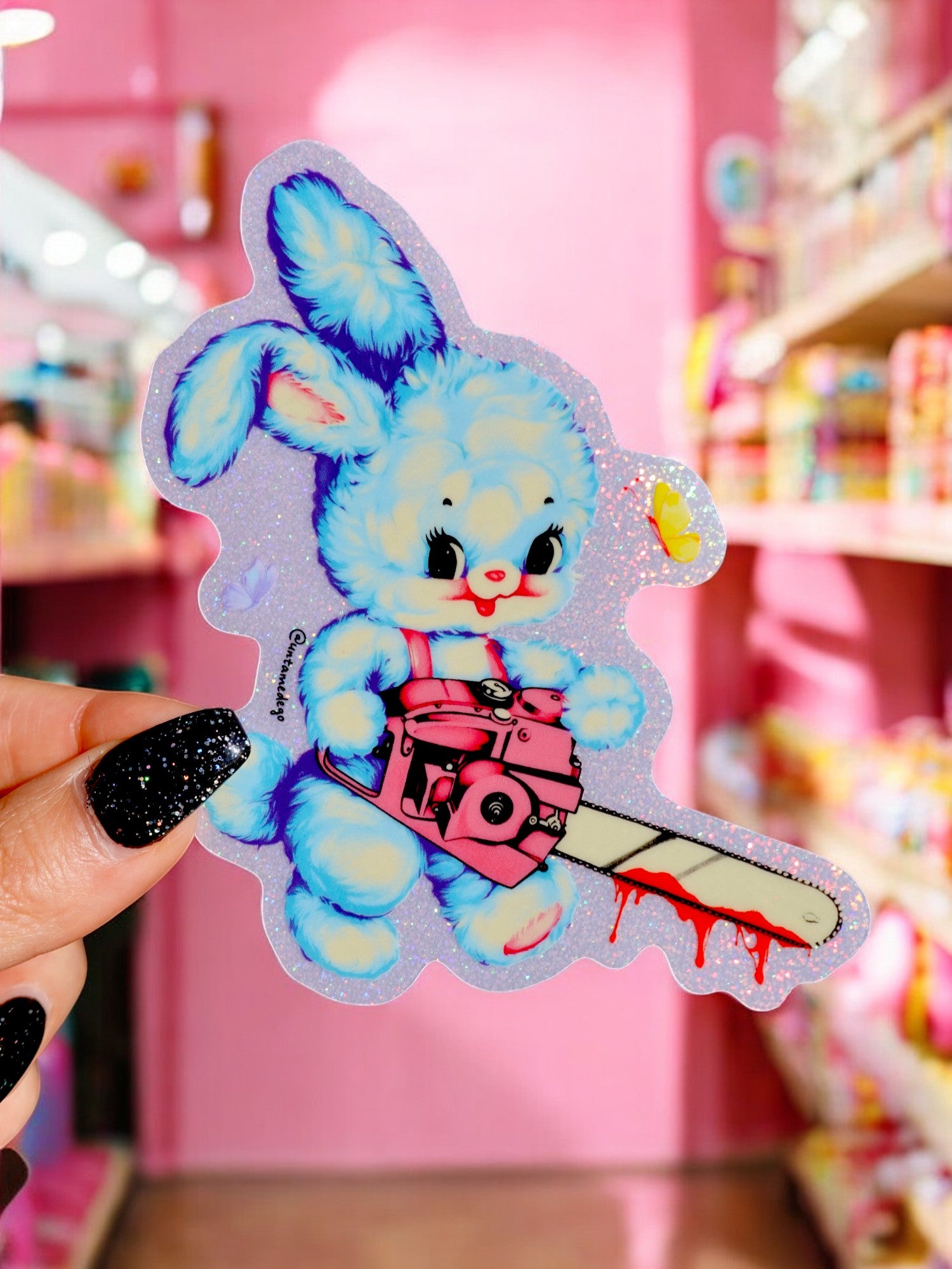 A sticker depicting a cartoon image of a blue rabbit holding a pink chainsaw with a glittery finish, displayed in front of a pink background.