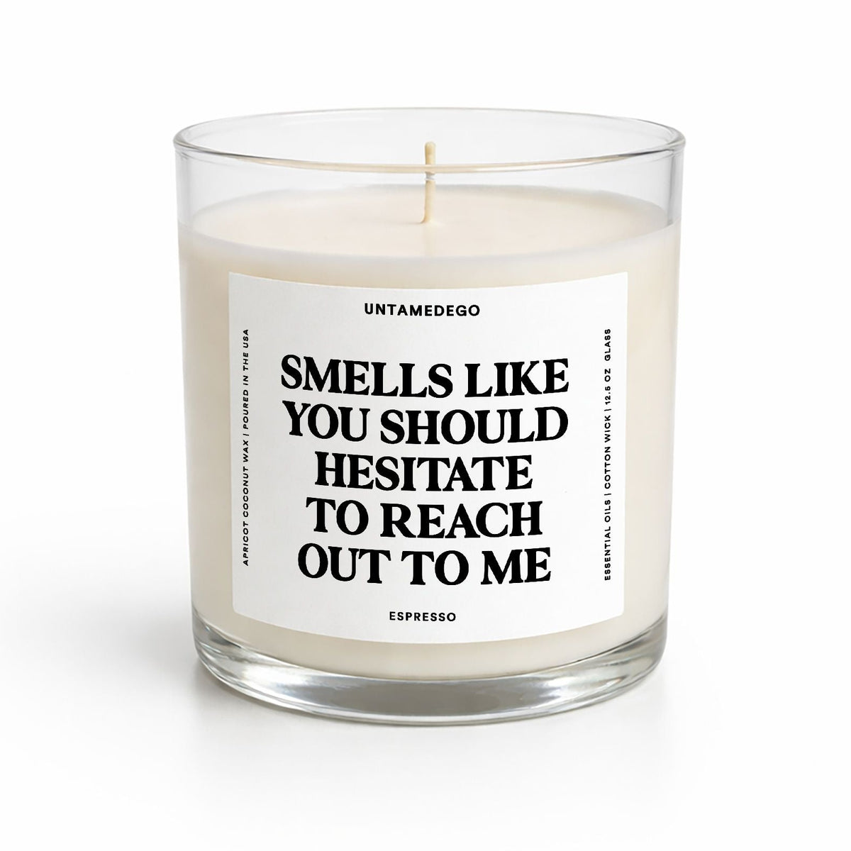 A clear glass jar candle with white wax and a white label featuring bold black text that reads, 'SMELLS LIKE YOU SHOULD HESITATE TO REACH OUT TO ME.' The brand name 'UNTAMEDEGO' is visible at the top of the label, with 'ESPRESSO' at the bottom.