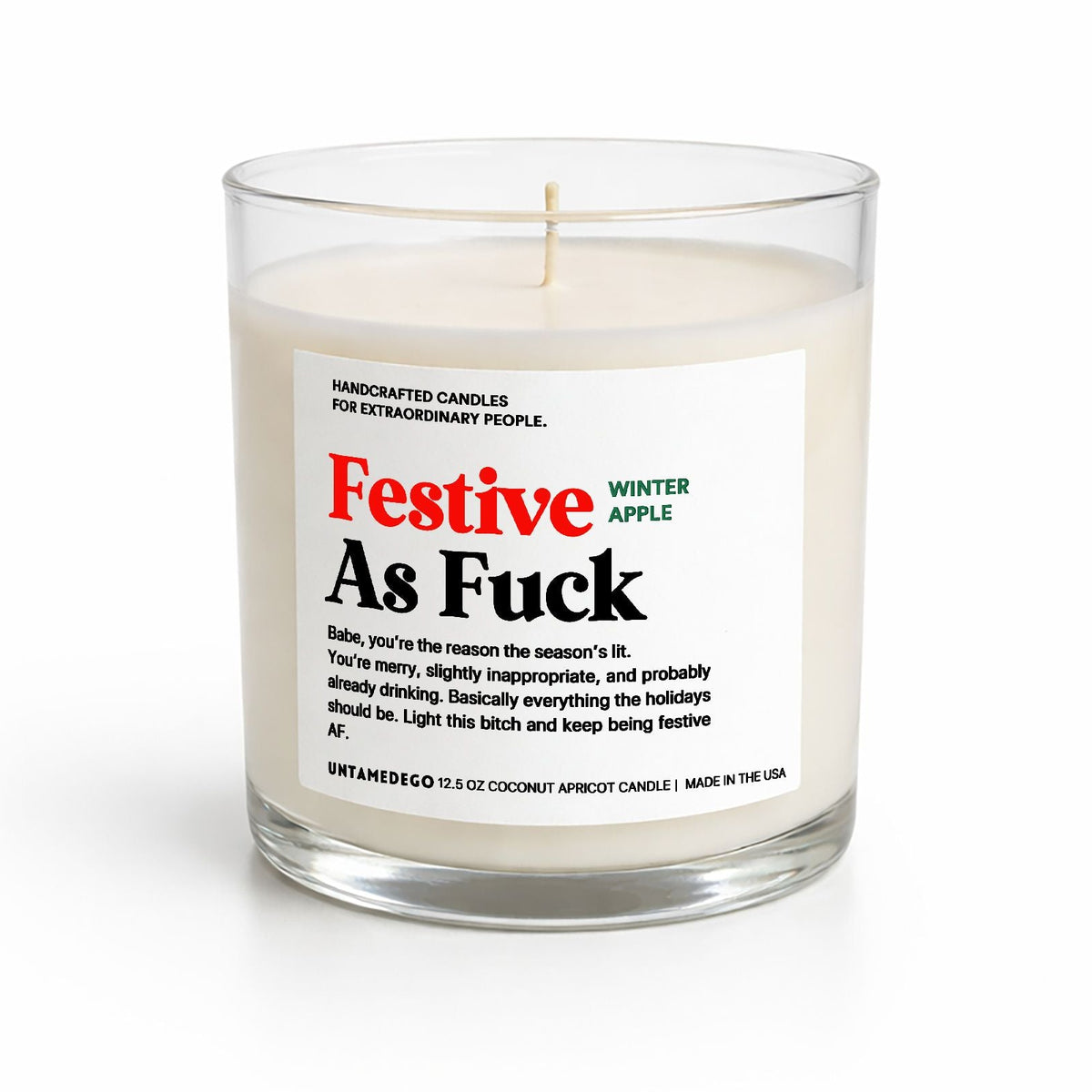 A 12 oz coconut apricot candle in a clear glass jar with a white label that reads 'Festive As Fuck' by Fraser Fir. The candle is lit, showing a soft warm glow, and is photographed on a clean white background.