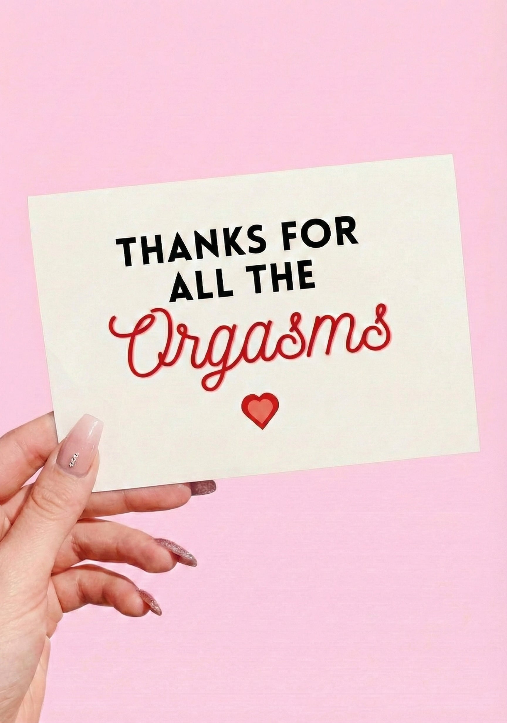 Greeting card with black and red text and a heart graphic on a light pink background, photographed being held by a hand