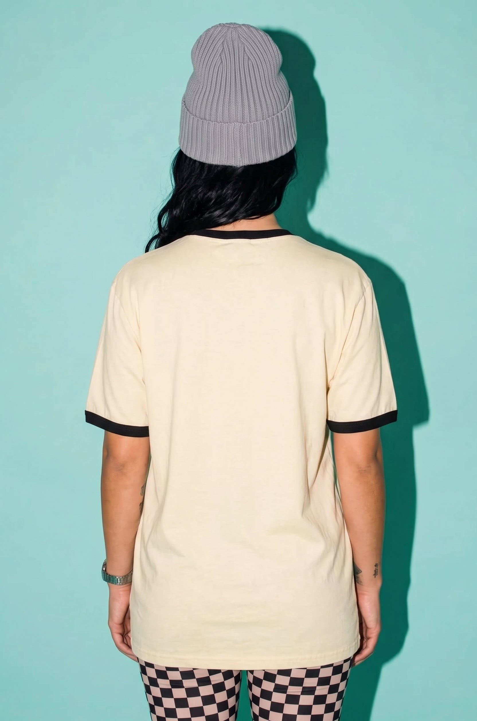A person shown from the back wearing a cream-colored ringer t-shirt with black trim on the collar and sleeve cuffs, paired with a gray ribbed beanie and black-and-white checkered leggings, set against a solid teal background.