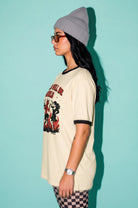 A woman in profile wearing a cream ringer t-shirt with a red and black graphic print, a gray beanie, and glasses, standing against a solid light teal background.