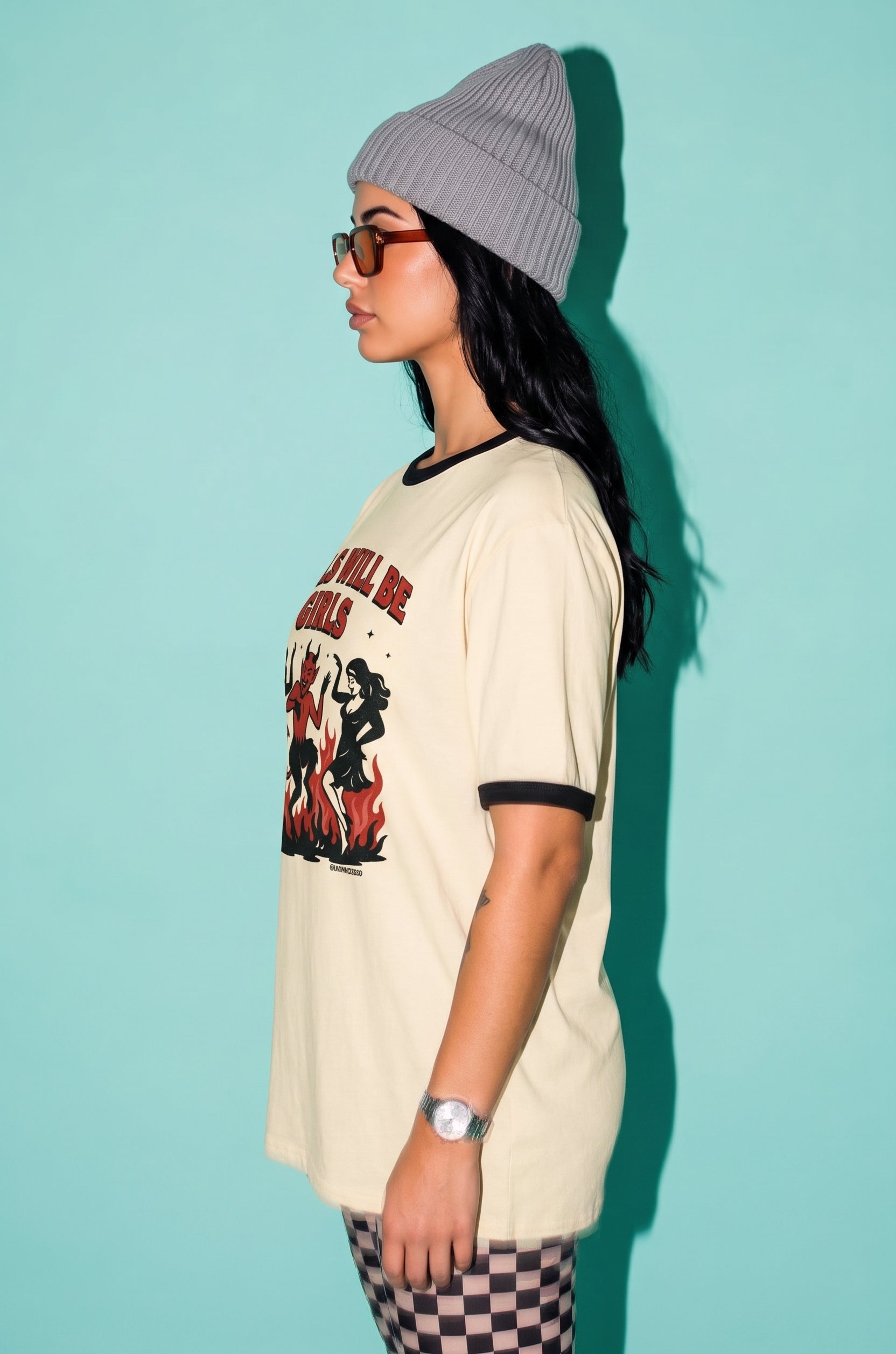 A woman in profile wearing a cream ringer t-shirt with a red and black graphic print, a gray beanie, and glasses, standing against a solid light teal background.
