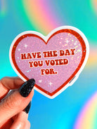 Heart-shaped sticker with 'Have the day you voted for.' text held by a hand against a colorful background