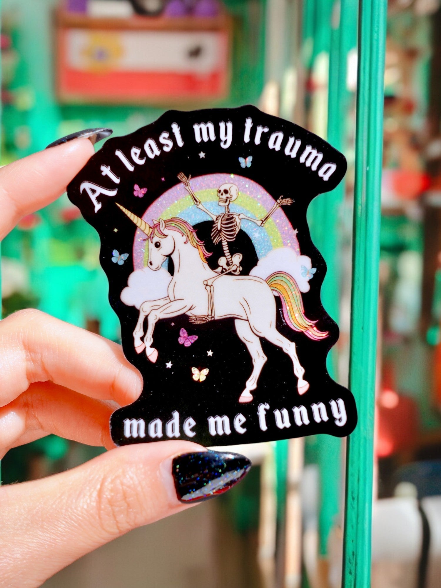 Holographic glitter vinyl sticker showing a skeleton in a party hat riding a unicorn under a rainbow with butterflies and clouds. Bold white text reads At Least My Trauma Made Me Funny.