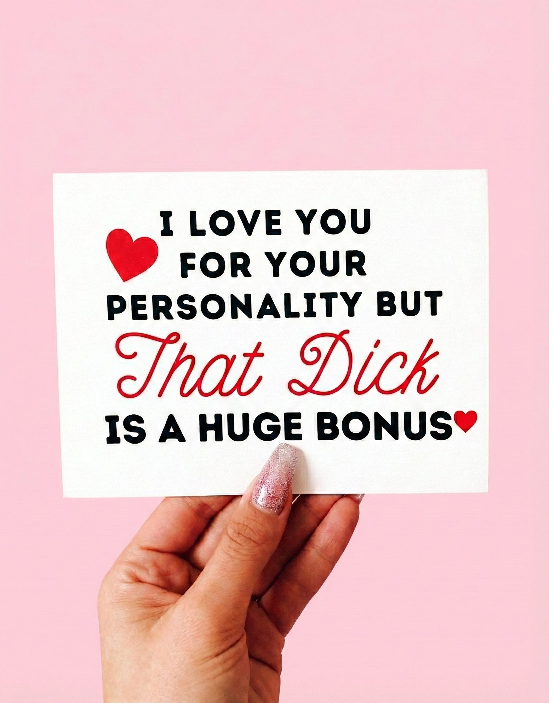 Greeting card displaying a humorous adult phrase in black and red text with heart graphics, photographed being held by a hand against a pink background