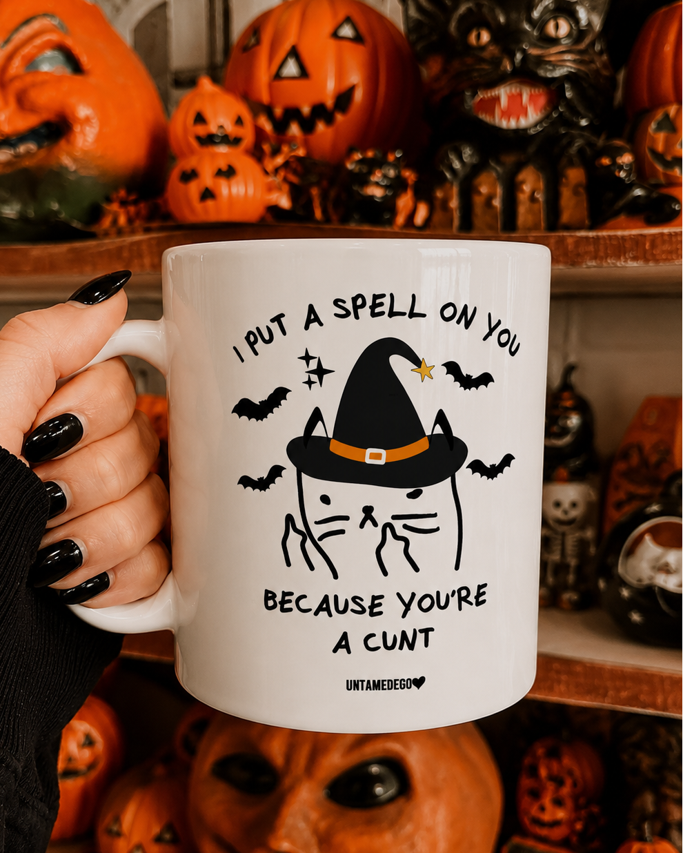 A hand with black nail polish holds a white ceramic mug featuring a black and orange witch cat graphic and the text 'I PUT A SPELL ON YOU BECAUSE YOU'RE A CUNT'. The background shows shelves filled with Halloween decorations, including carved pumpkins and spooky figurines.