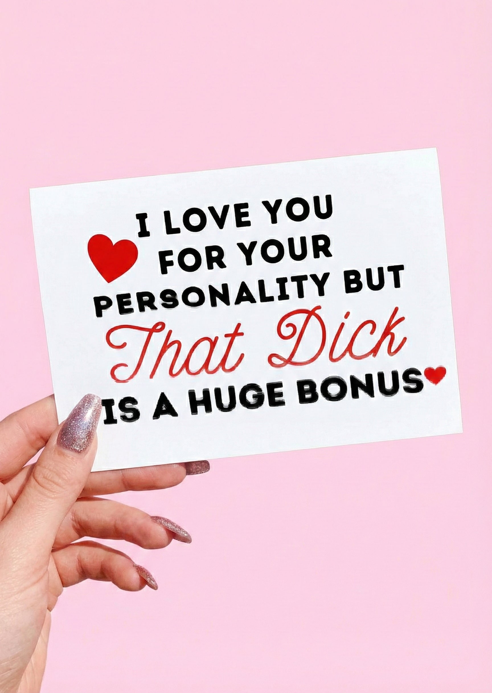 Greeting card displaying a humorous adult phrase in black and red text with heart graphics, photographed being held by a hand against a pink background