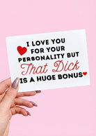 Greeting card displaying a humorous adult phrase in black and red text with heart graphics, photographed being held by a hand against a pink background