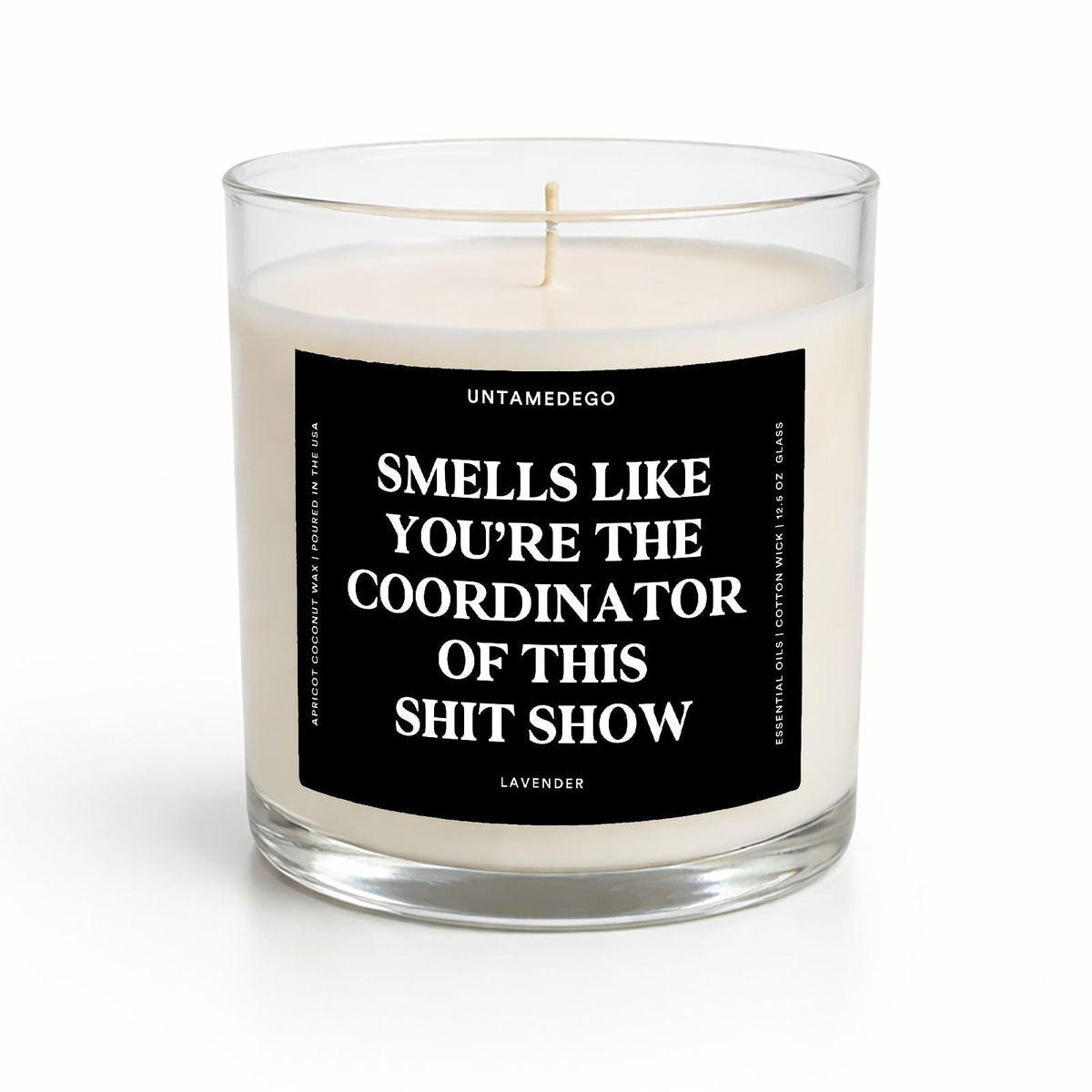 A clear glass jar candle with white wax and a black label featuring bold white text that reads, 'SMELLS LIKE YOU'RE THE COORDINATOR OF THIS SHIT SHOW.' The label also shows the brand name 'UNTAMEDEGO' and the scent 'LAVENDER.