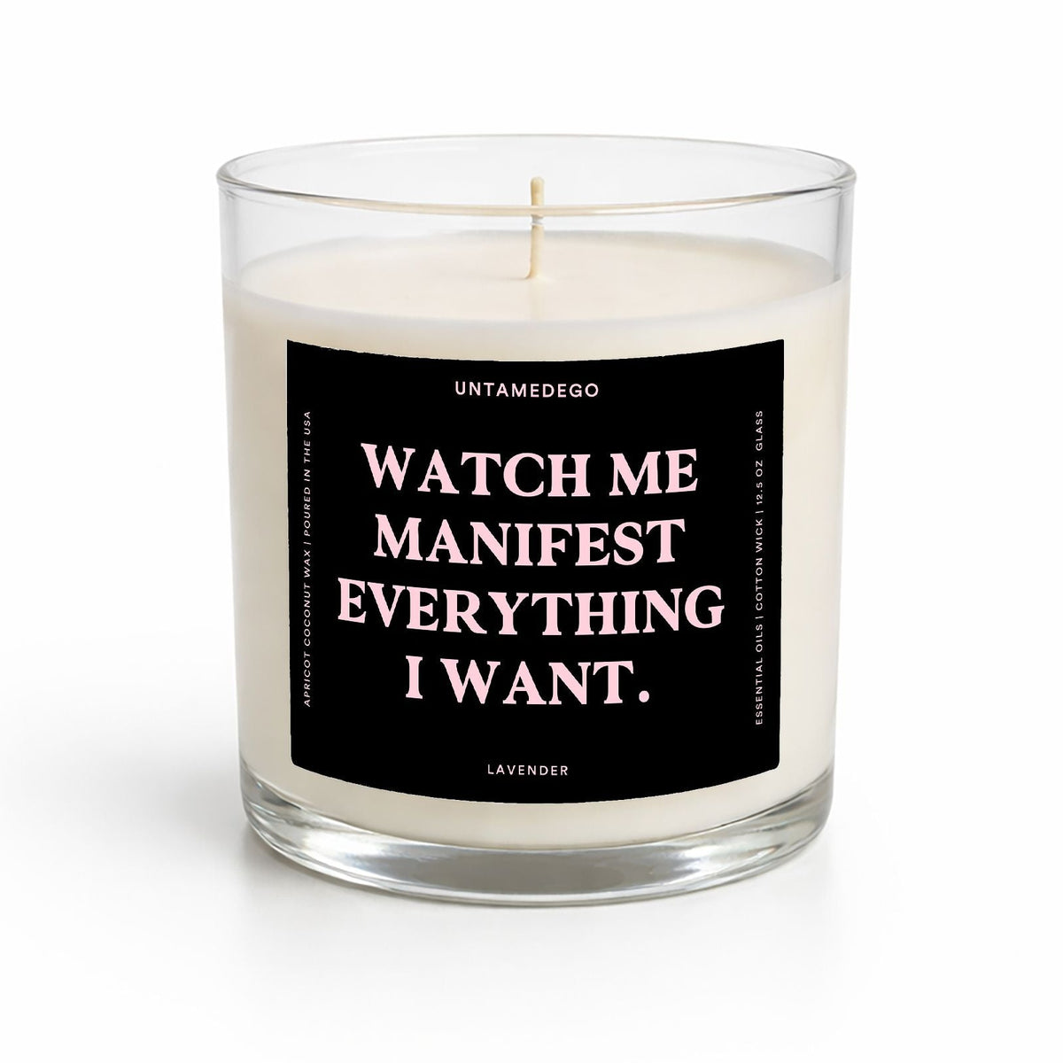 A clear glass jar candle with white wax and a black label featuring the text 'WATCH ME MANIFEST EVERYTHING I WANT.' in white and pink lettering, labeled 'UNTAMEDEGO' and 'LAVENDER' at the bottom.