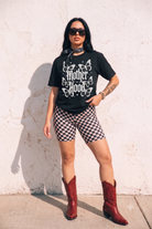 A woman stands against a white stucco wall wearing a black graphic T-shirt with white 'Mother Hood' text and butterfly artwork, black-and-white checkered biker shorts, a dark bandana at the neck, and red cowboy boots.