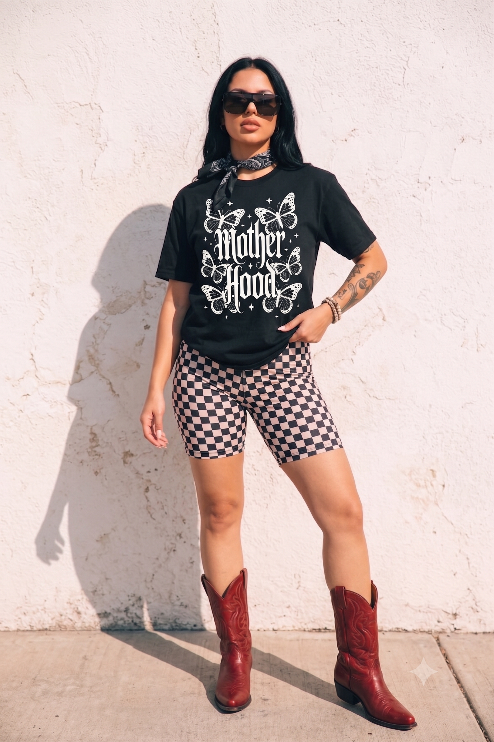 A woman stands against a white stucco wall wearing a black graphic T-shirt with white 'Mother Hood' text and butterfly artwork, black-and-white checkered biker shorts, a dark bandana at the neck, and red cowboy boots.