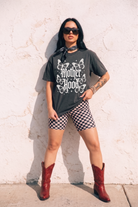 A woman stands against a textured white wall wearing a dark charcoal graphic T-shirt with 'Mother Hood' and butterfly artwork, black-and-white checkered biker shorts, and red cowboy boots.
