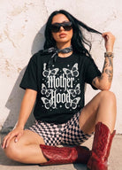 A woman wearing a black graphic t-shirt with 'Mother Hood' and butterflies, black-and-white checkered shorts, and red cowboy boots, sitting against a textured white wall.