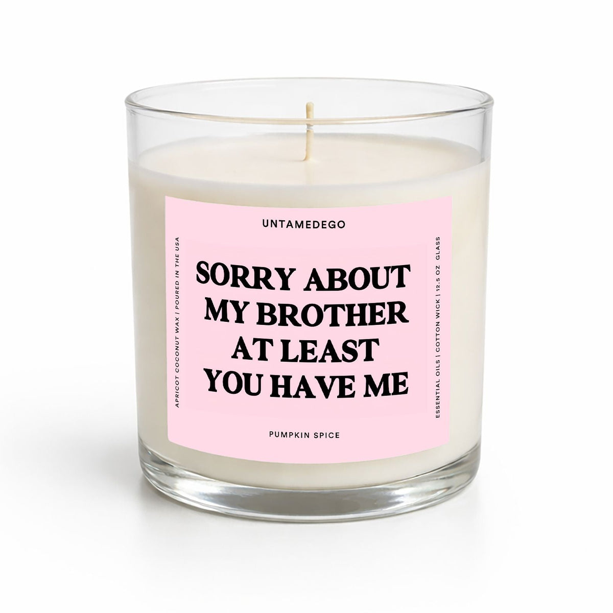 A single-wick candle in a clear glass jar with white wax and a pink label that reads 'SORRY ABOUT MY BROTHER AT LEAST YOU HAVE ME' by Untamedego.