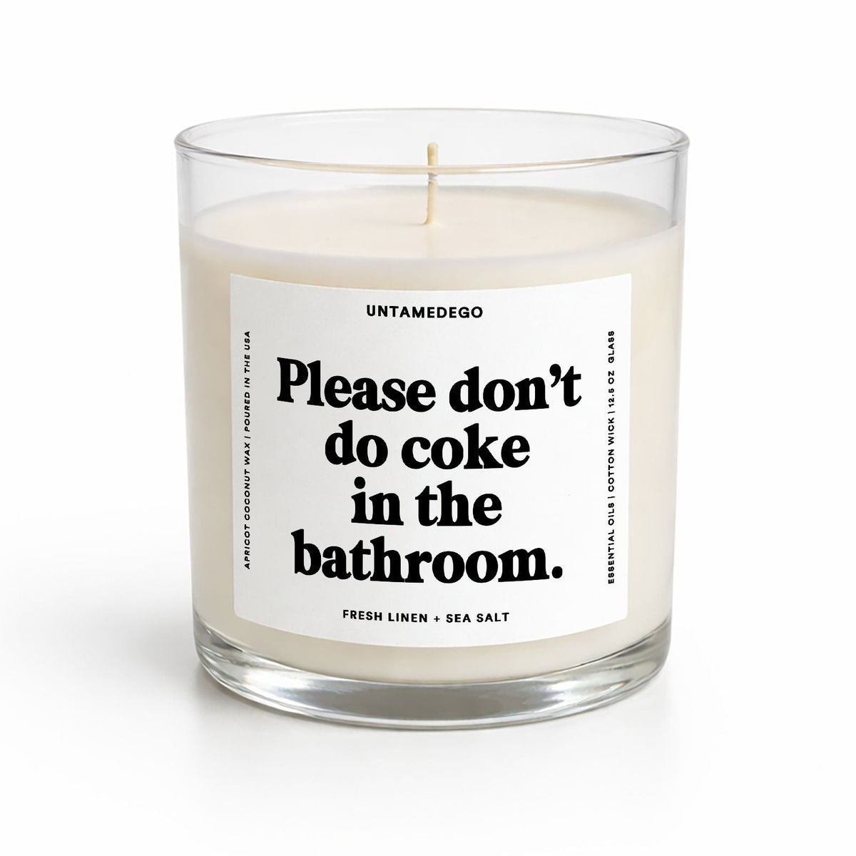 A clear glass jar candle with creamy off-white wax and a white label featuring bold black text that reads, 'Please don't do coke in the bathroom.' The brand name 'UNTAMEDEGO' is visible at the top of the label.