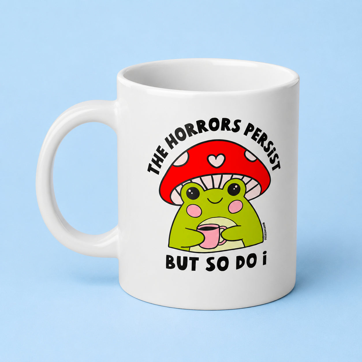 A white ceramic coffee mug featuring a cartoon graphic of a green frog under a red mushroom cap with the text 'THE HORRORS PERSIST BUT SO DO I', set against a light blue background.