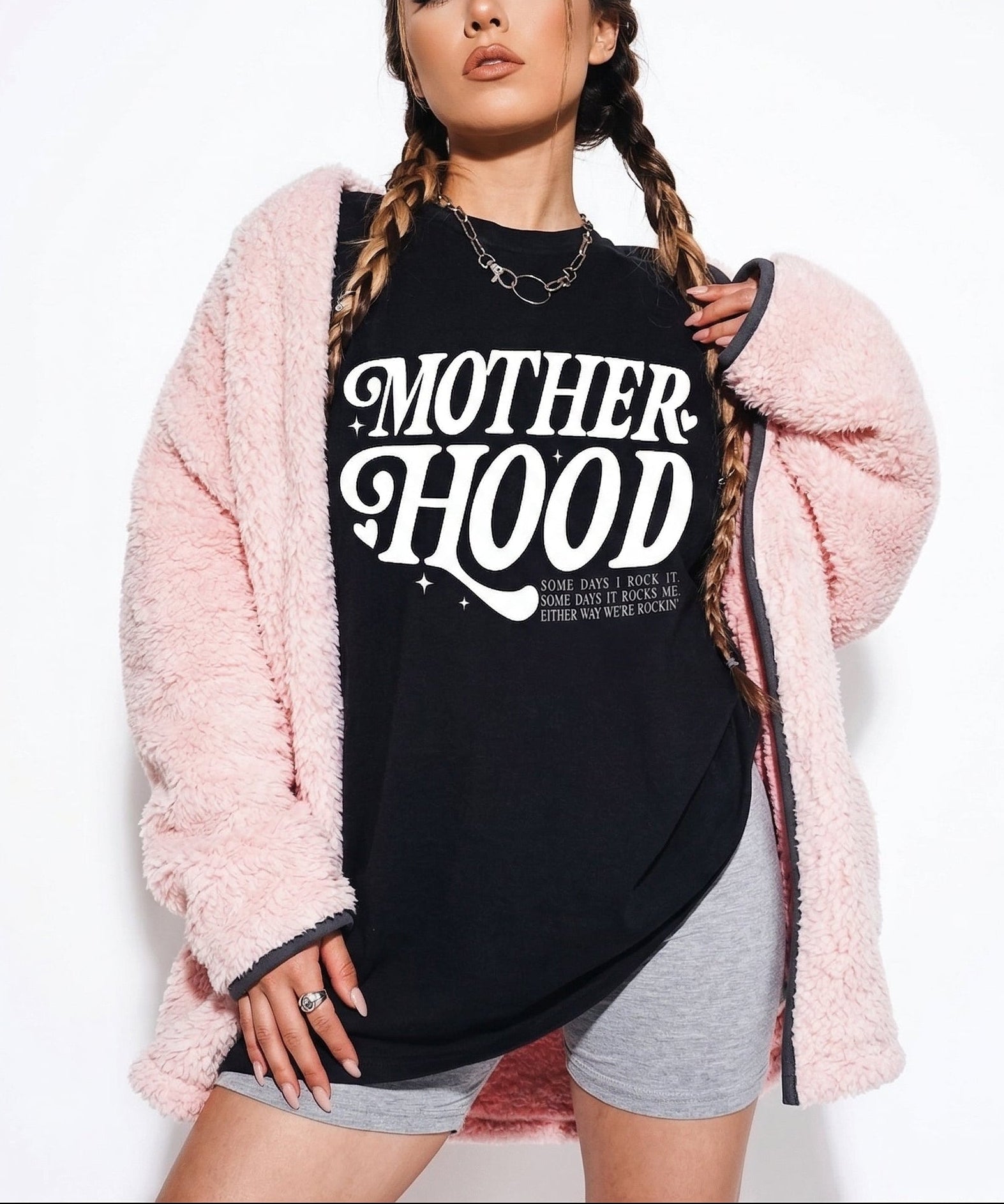 Woman wearing a black t-shirt with 'MOTHERHOOD' text, pink cardigan, and gray shorts on a white background