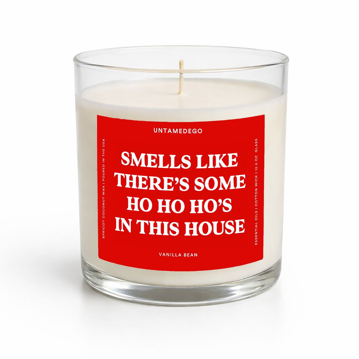 A clear glass jar candle with white wax and a prominent red label featuring white text that reads, 'SMELLS LIKE THERE'S SOME HO HO HO'S IN THIS HOUSE.' The brand name 'UNTAMEDEGO' is visible at the top of the label, and the scent 'VANILLA BEAN' is at the bottom.