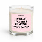 A single-wick candle in a clear glass jar with cream-colored wax and a light pink label that reads 'SMELLS LIKE SHE'S READING SMUT AGAIN' in bold black text. The brand name 'UNTAMEDEGO' is visible at the top of the label.