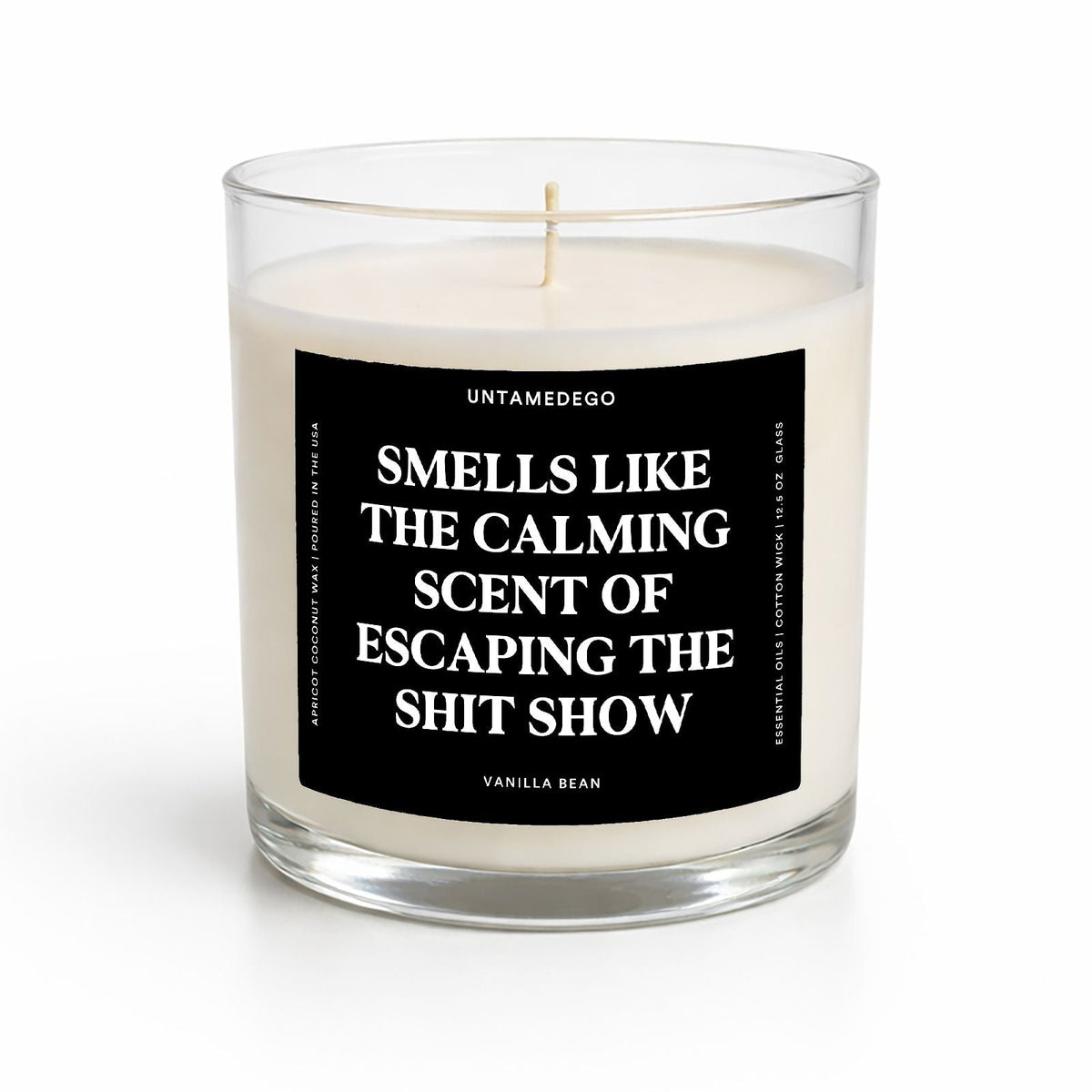 A clear glass jar candle with white wax and a black label featuring bold white text that reads, 'SMELLS LIKE THE CALMING SCENT OF ESCAPING THE SHIT SHOW.' The brand name 'UNTAMEDEGO' is visible at the top of the label.