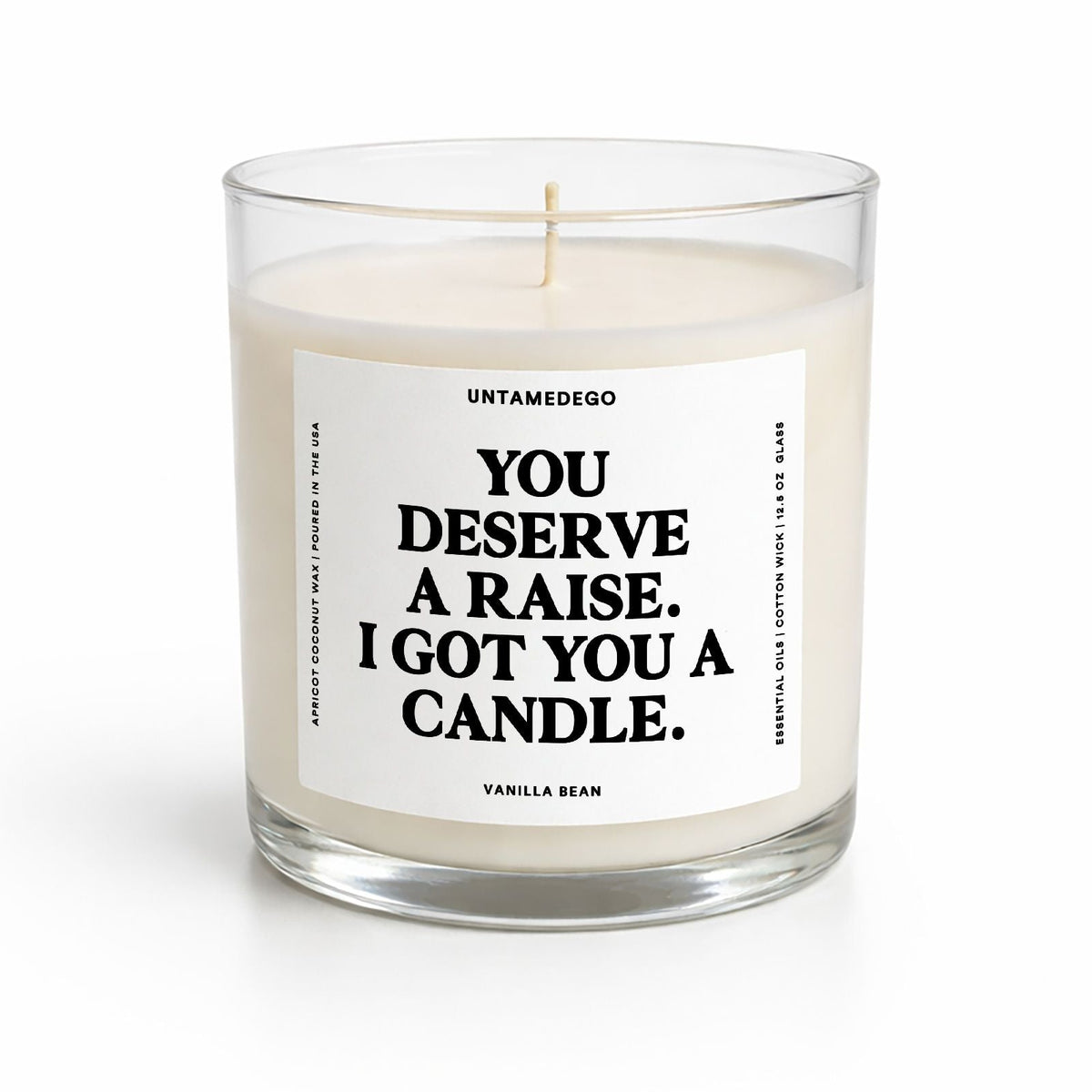 A single-wick candle in a clear glass jar with cream-colored wax and a white label featuring black text that reads, 'YOU DESERVE A RAISE. I GOT YOU A CANDLE.