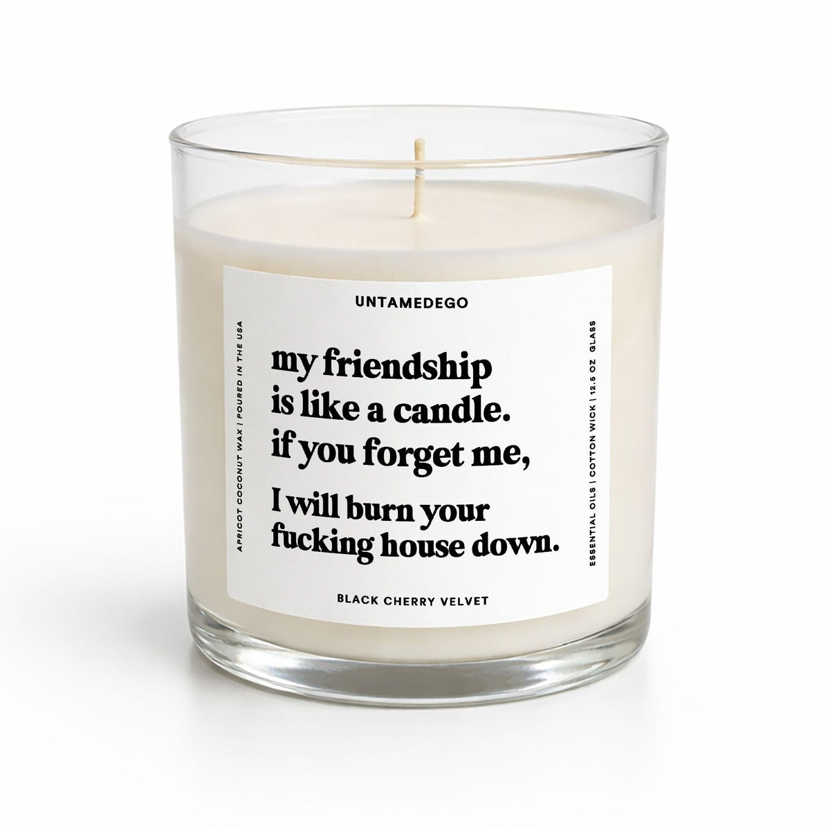 A clear glass jar candle with cream-colored wax and a white label featuring black text. The label reads 'UNTAMEDEGO' and 'my friendship is like a candle, if you forget me, I will burn your fucking house down.