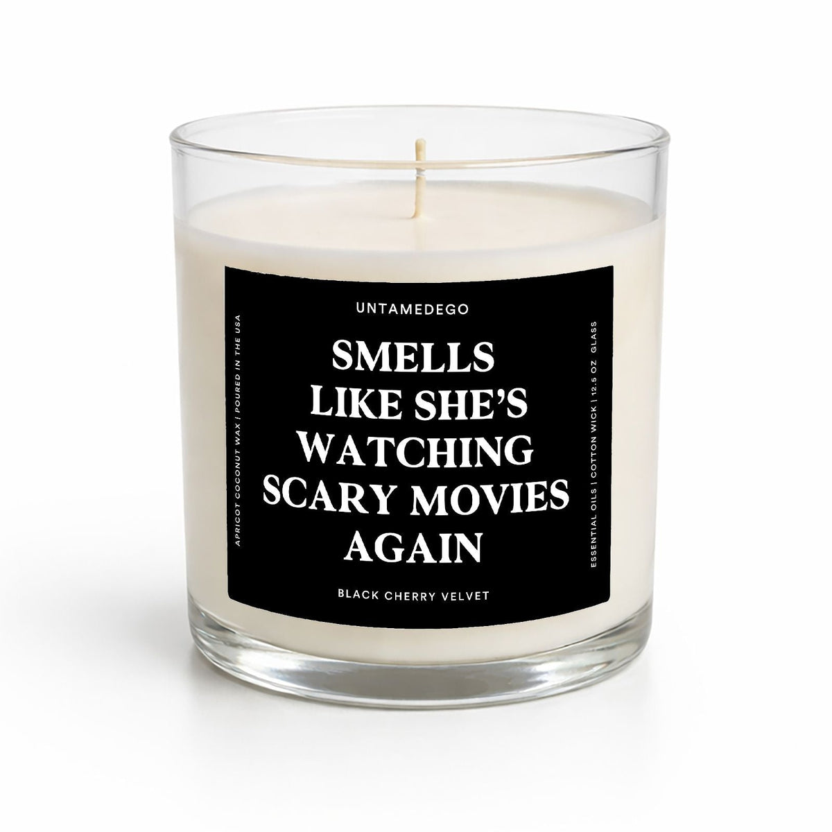 A clear glass jar candle with white wax and a black label featuring white text that reads 'SMELLS LIKE SHE'S WATCHING SCARY MOVIES AGAIN' and the brand name 'UNTAMEDEGO'.