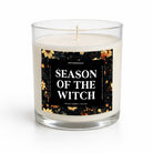 A three-quarter studio shot of a candle in a clear glass jar on a white background. The candle has off-white wax and a black label with a floral border and white text reading 'SEASON OF THE WITCH' and 'UNYAMBIOSO'.